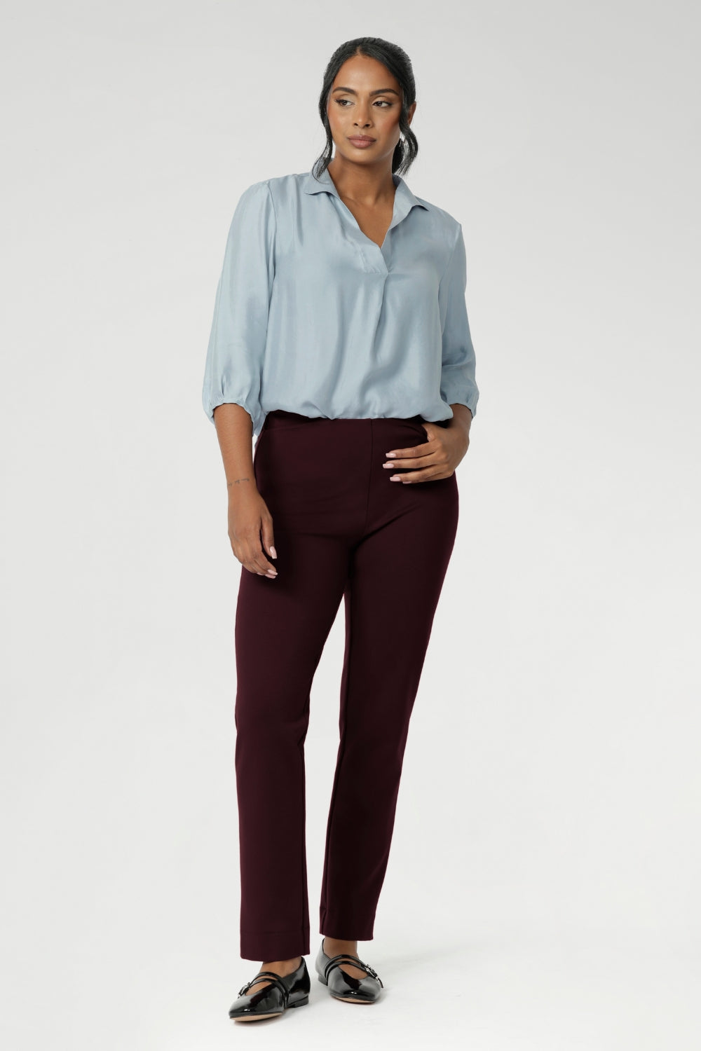 Semi-fitted through the body, it features a classic collar neckline and subtle tuck details below the back neckline, adding gentle structure without compromising comfort. Over 30s woman wears her light blue shirt tucked into burgundy coloured work pants.