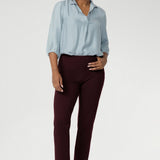 Semi-fitted through the body, it features a classic collar neckline and subtle tuck details below the back neckline, adding gentle structure without compromising comfort. Over 30s woman wears her light blue shirt tucked into burgundy coloured work pants.