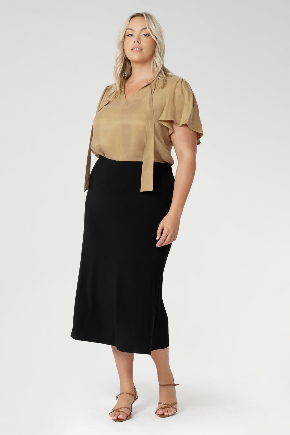 plus size woman wears black sleek skirt with streamlined design that can easily be dressed up for a more elevated look or worn casually - making it a truly versatile wardrobe staple. Model pairs black midid skirt with plain white top and a silver sneaker.