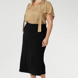 plus size woman wears black sleek skirt with streamlined design that can easily be dressed up for a more elevated look or worn casually - making it a truly versatile wardrobe staple. Model pairs black midid skirt with plain white top and a silver sneaker.