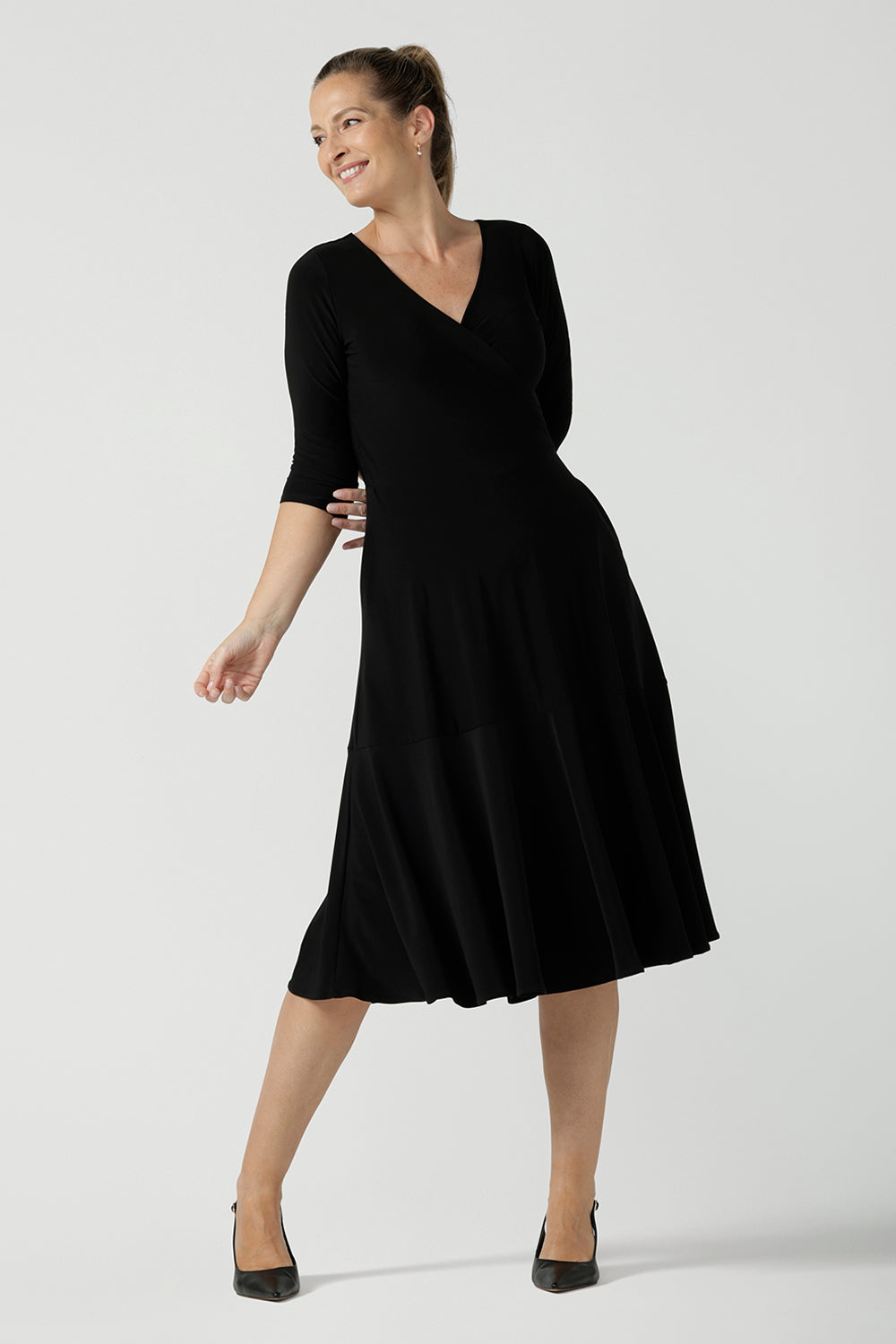 Size 10 woman wears the Bettina dress in black soft jersey. Wrap front reversible design with pockets and a tier. Styled back with black sling back shoes. Made in Australia for women size 8 - 24.