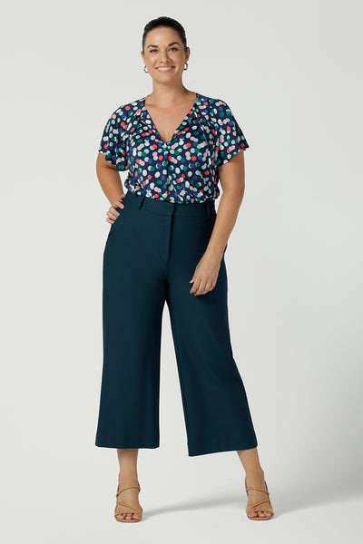 A size 12 happy woman wears a Bowie top with a flutter sleeve and v-neck with neck ties. All over polka dot print on a soft slinky jersey. Styled back with a Yael Pant in petrol, a high waist tailored work pant. Comfortable work top for women. Made in Australia size 8 - 24.