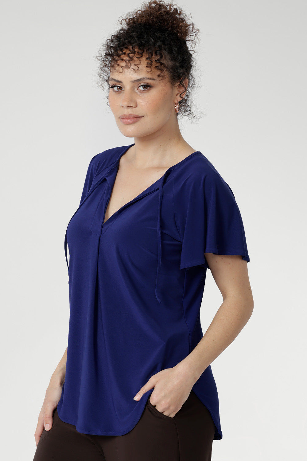 The Bowie Top in Sapphire is designed for comfort and confidence. Made from dry-touch jersey with soft stretch, it drapes beautifully in a relaxed fit with a flattering V-neckline, pleated front, and playful tie accents. Flutter sleeves and a shirttail hem add the perfect finishing touch for workdays or downtime alike.