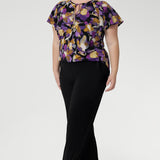 The vibrant painterly-style floral print, bursting with rich purples and soft yellow hues, brings a fresh pop of colour to any outfit. Plus size model wears her printed blouse with flutter sleeved with black wide leg pants and heels.
