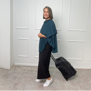 Dress for travel in this stylish travel outfit by Australia and New Zealand women's clothing brand, Leina & Fleur. An over 50 woman wears a petrol green jersey with black wide leg travel pants. 