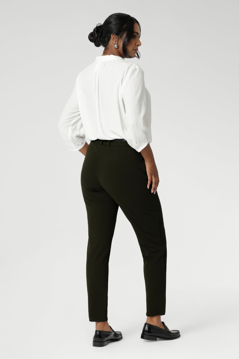 Back view image of The Corbin Pant in Olive worn with lightweight blouse and black loafers. The pants are designed with angled front pockets, giving them a modern touch, while the back darts ensure a tailored fit that contours to the shape of the body.