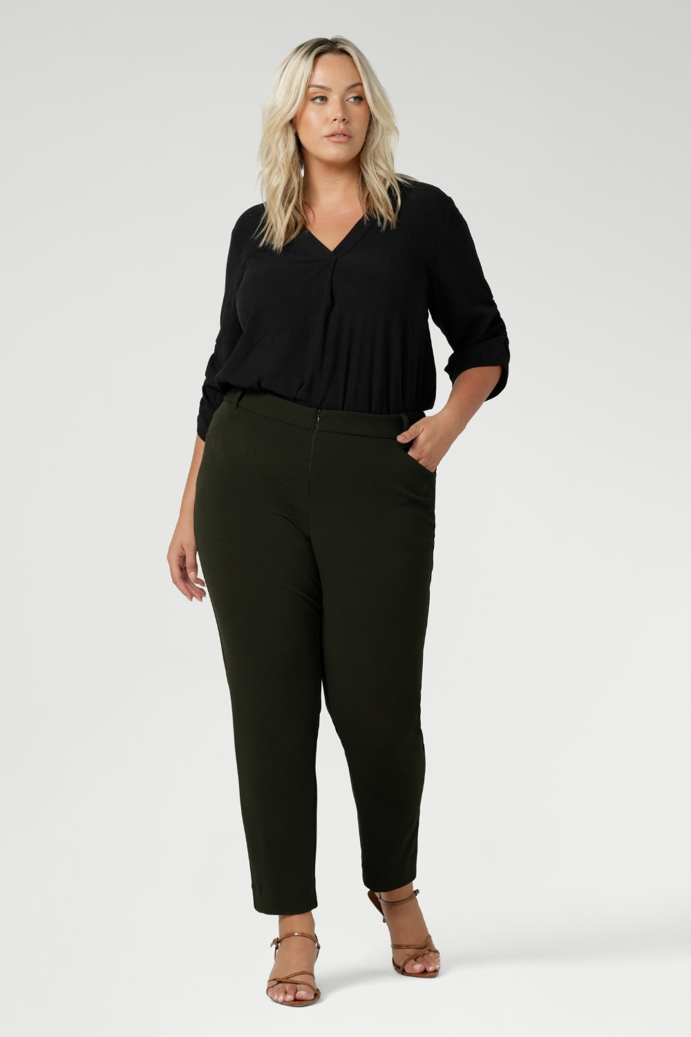 Plus size woman wears her Corbin Pants in olive with lightweight black blouse and brown kitten heels. The pants are designed with angled front pockets, giving them a modern touch, while the back darts ensure a tailored fit that contours to the shape of the body. Available in sizes 8-24.