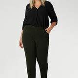 Plus size woman wears her Corbin Pants in olive with lightweight black blouse and brown kitten heels. The pants are designed with angled front pockets, giving them a modern touch, while the back darts ensure a tailored fit that contours to the shape of the body. Available in sizes 8-24.
