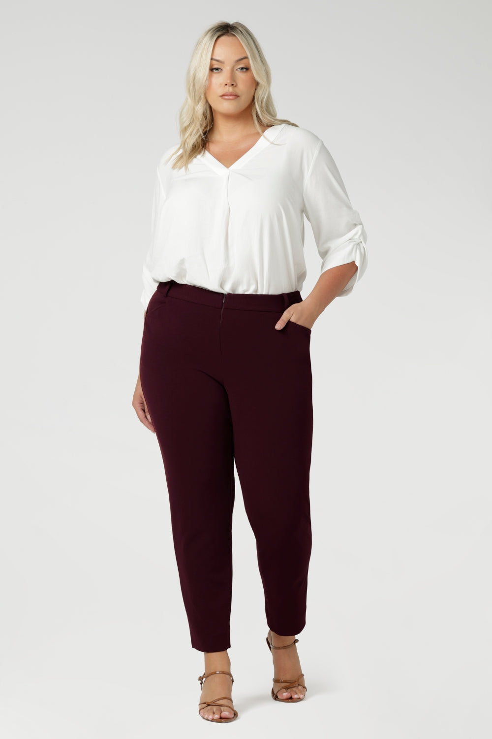 Designed with a mid-rise fit, they feature a waistband with belt loops for added styling options. The invisible front zip with an internal button ensures a sleek, clean look while providing a secure fit. Size 18 woman wears her Corbin Pants with white blouse and brown heels.
