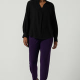 A size 10 woman wearing the Indi Pant in Amethyst. An Indi pant in amethyst with a mid rise waistband, tapered ankle balloon leg style. A made in Australia pant for women. Perfect travel friendly non-crease pant. Size 8-10. Styled back with a black Ellery Shirt in Tencel.