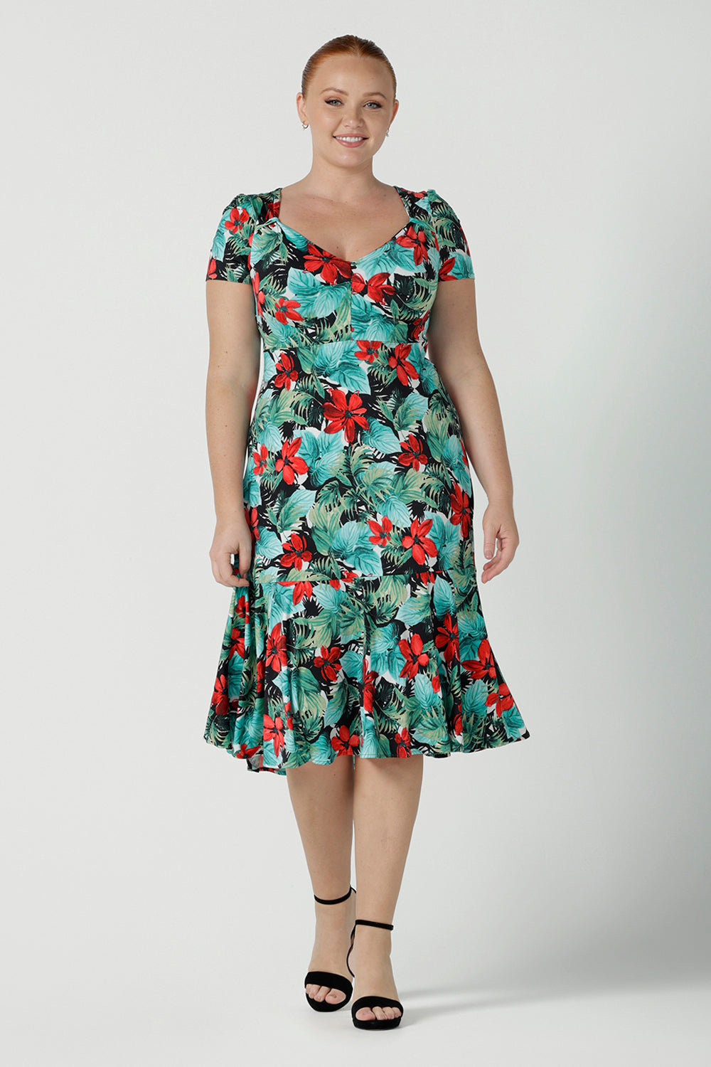 Size 12  woman wears the Jillian dress in Havana. A beautiful tropical print with a green leaf print and red flower on a white base that is tropical inspired. A sweetheart neckline style with an empire line. Tier on hem. Made in Australia for women. Size 8 - 24.