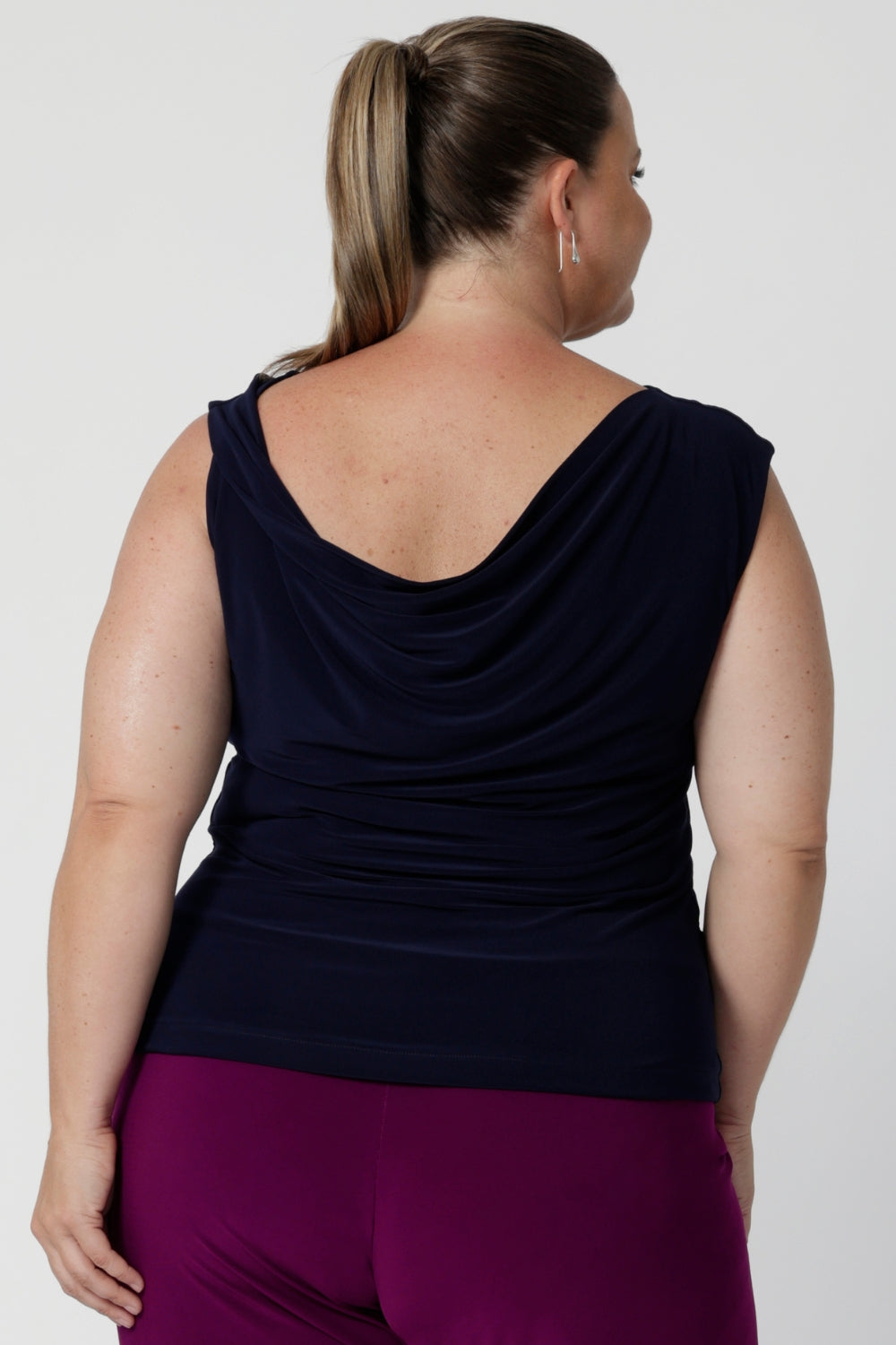 Reversible navy top with a cowl neckline and twisted shoulder detail facing the back for a feminine drape across the back and shoulders. Plus size woman wears her navy workwear top with magenta coloured pants.