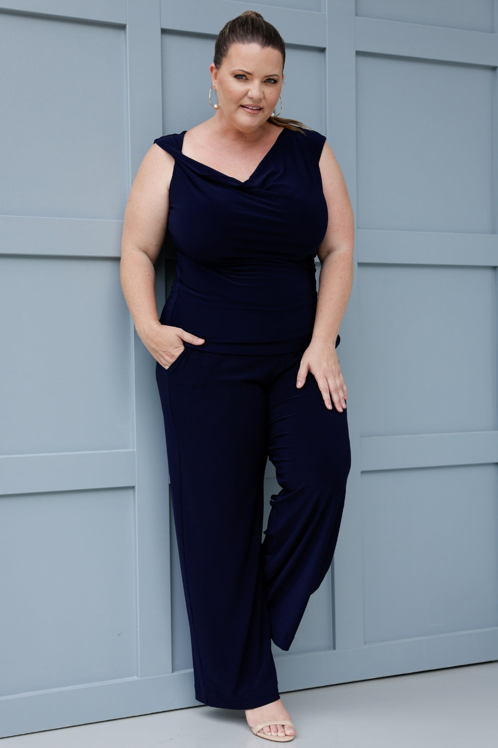 Image features woman over 40 wearing a navy cowl neckline top, paired with matching navy pants for a chic faux jumpsuit effect. The reversible double-layer design allows you to wear it with the cowl neck facing forward, or flip it around to reveal a boat-neck style, offering a more open, structured silhouette. Shop Jordan Tops now in sizes 8-24.