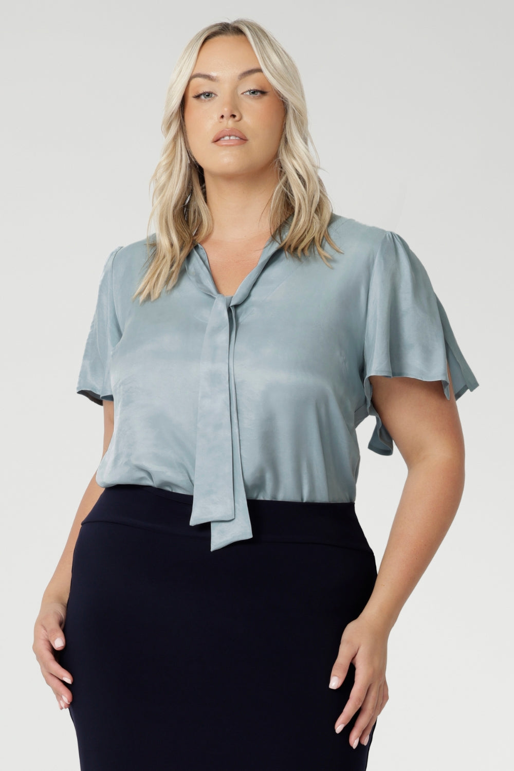 Image of plus size model wearing suede touch blouse styled with navy work skirt and silver accessories. The V-neckline with adjustable neckties allows for a customisable fit, adding a feminine touch to the look. Short flutter sleeves with slits at the shoulders create a breezy, modern feel, bringing a subtle hint of playful elegance to the piece.