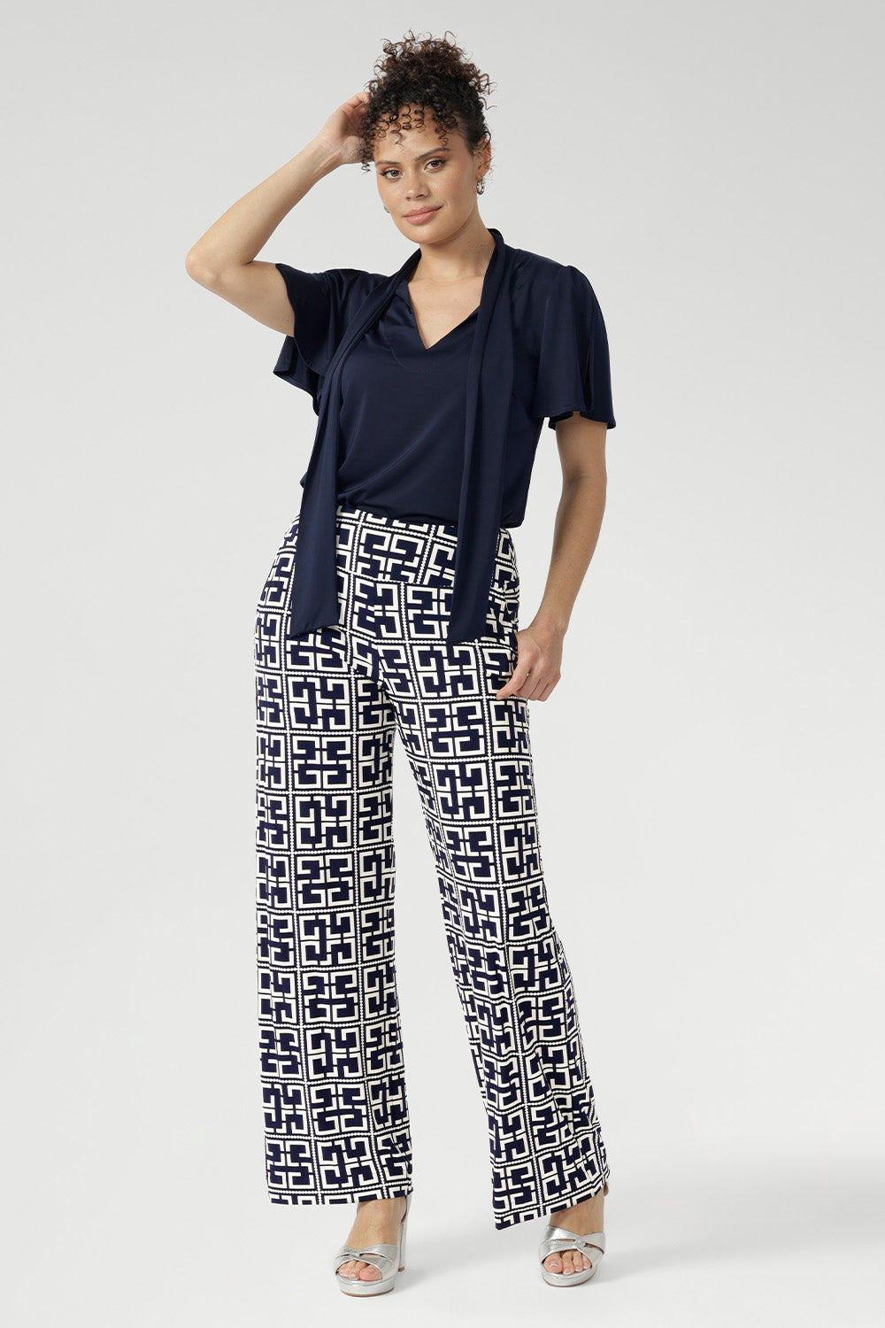 Full body front view of 30 year old woman wearing a navy top tucked into printed pants. This short sleeved shirt has neck ties that can be styled in a bow or left untied. Easy care dry touch jersey fabric was used to create this garment.