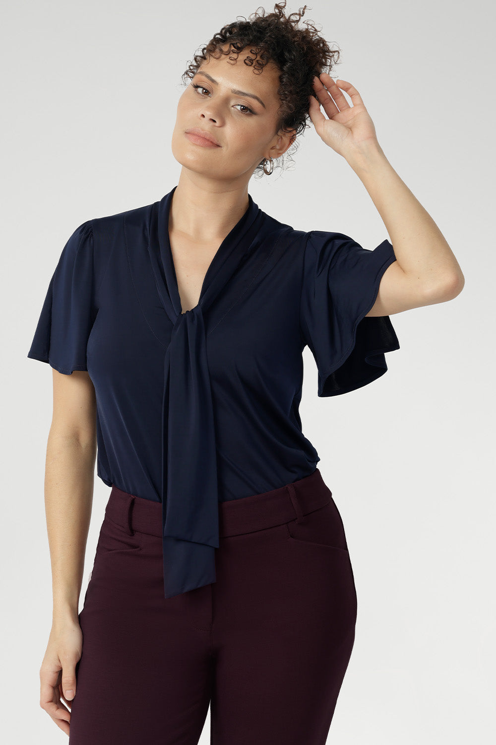 Woman wearing a navy blue top with neck ties detail and wine straight leg pants. Close up shot of 30 year old woman wears navy work shirt with neck ties that can be styled in many different ways. This work shirt is perfect for a corporate look and pairs well with most Leina + Fleur core pieces.