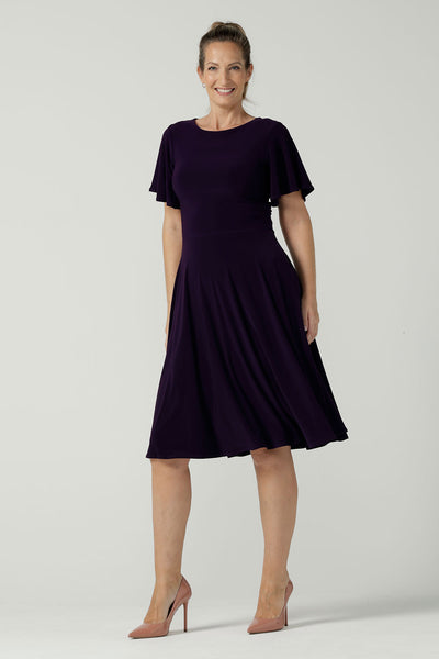 A size 10 woman wears the Monique dress in Amethyst. Jersey dress with flutter sleeve and rounded neckline. Full circle skirt and pockets. Made in Australia for women size 8 - 24.
