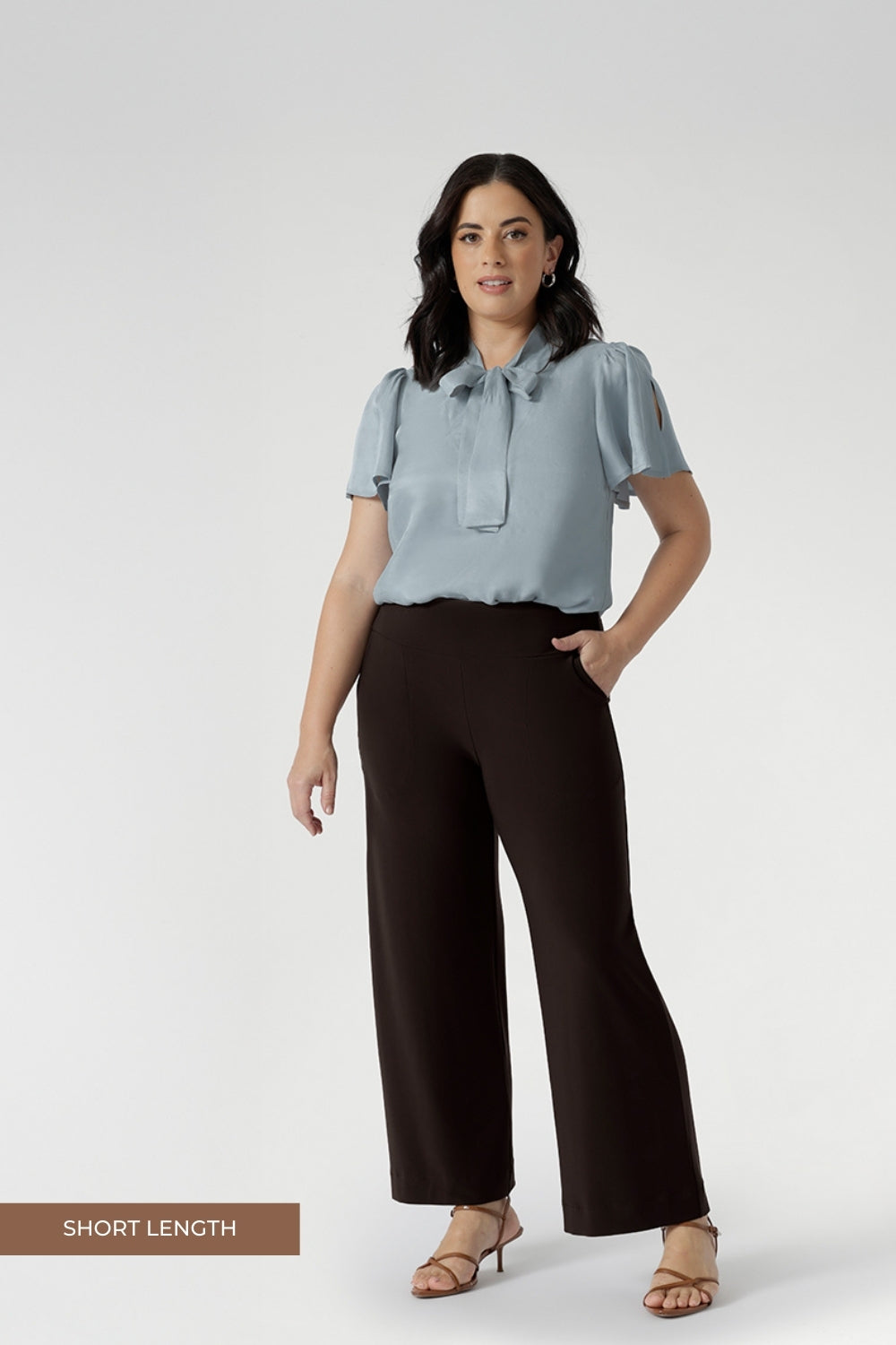 Petite height woman wears Monroe pants in cocoa in the short length. She pairs full lengths pants with a woman's blouse with neck ties. The pant includes a deep, double-layer waistband, side pockets with topstitching detail, a straight-leg cut, and a deep hem for added structure.