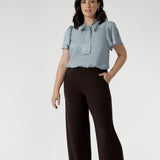 Petite height woman wears Monroe pants in cocoa in the short length. She pairs full lengths pants with a woman's blouse with neck ties. The pant includes a deep, double-layer waistband, side pockets with topstitching detail, a straight-leg cut, and a deep hem for added structure.