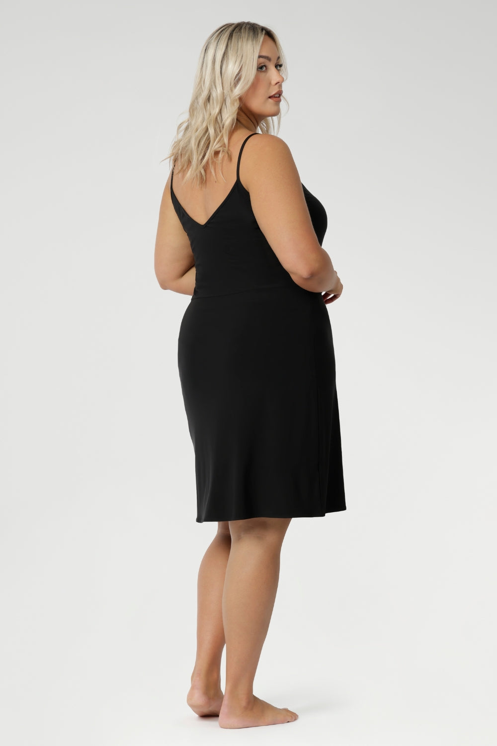 A size 18 woman wears a mini-length, reversible slip in black jersey fabric. The slip has thin straps and is worn with a V neck to the front. Made in Australia to layer under dresses and separates. Plus size slip dress for women. Available in size 8-24.