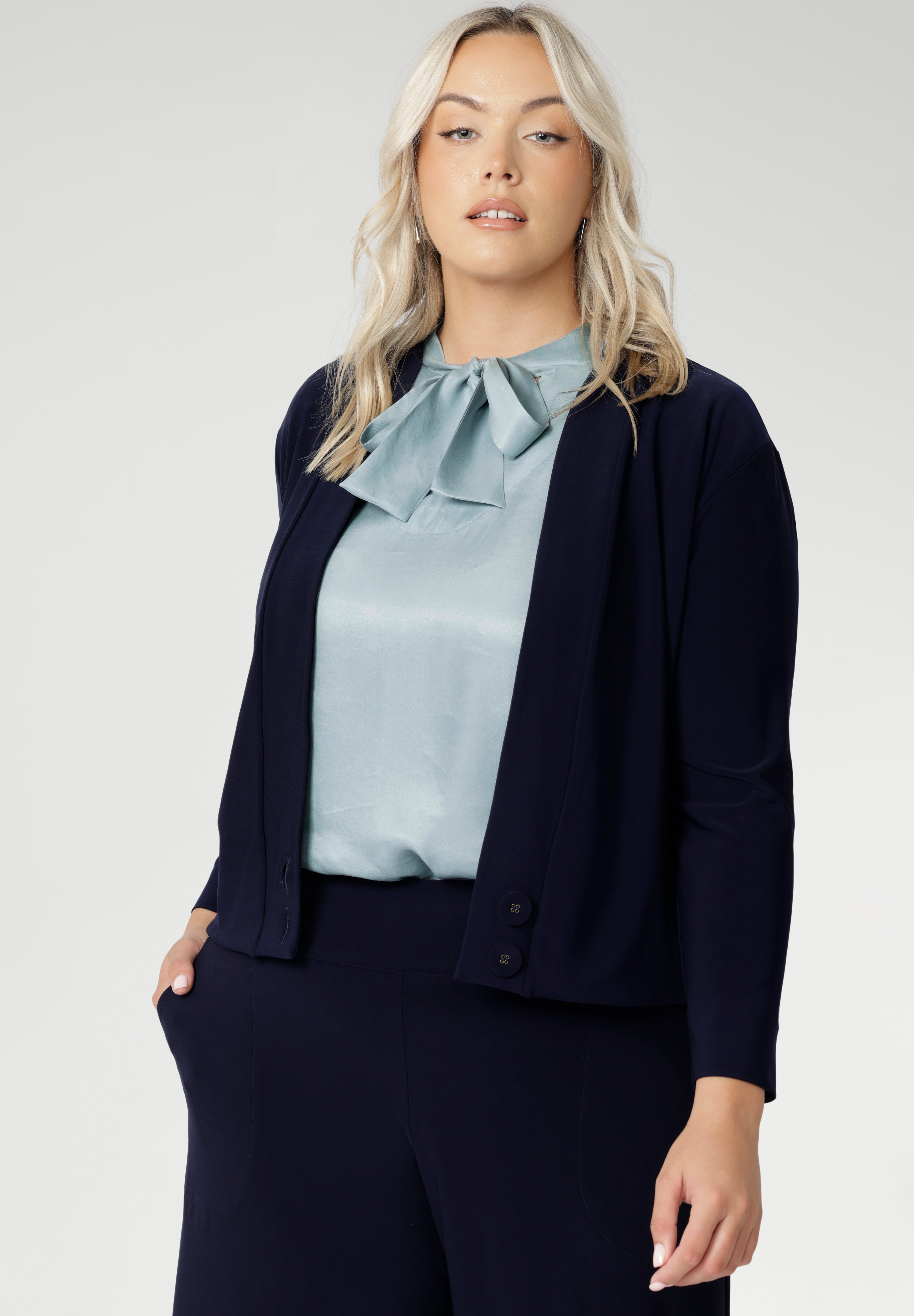 Plus size woman wears navy cardigan paired with woman's navy work pants and blue blouse with neck ties. The Jacardi features long sleeves, this jacket-cardigan hybrid is the perfect trans-seasonal cover up for women. Move into the cooler months in style, with this comfortable jacket for workwear and casual layering. Available in sizes 8-24.