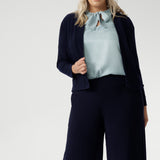 Plus size woman wears navy cardigan paired with woman's navy work pants and blue blouse with neck ties. The Jacardi features long sleeves, this jacket-cardigan hybrid is the perfect trans-seasonal cover up for women. Move into the cooler months in style, with this comfortable jacket for workwear and casual layering. Available in sizes 8-24.