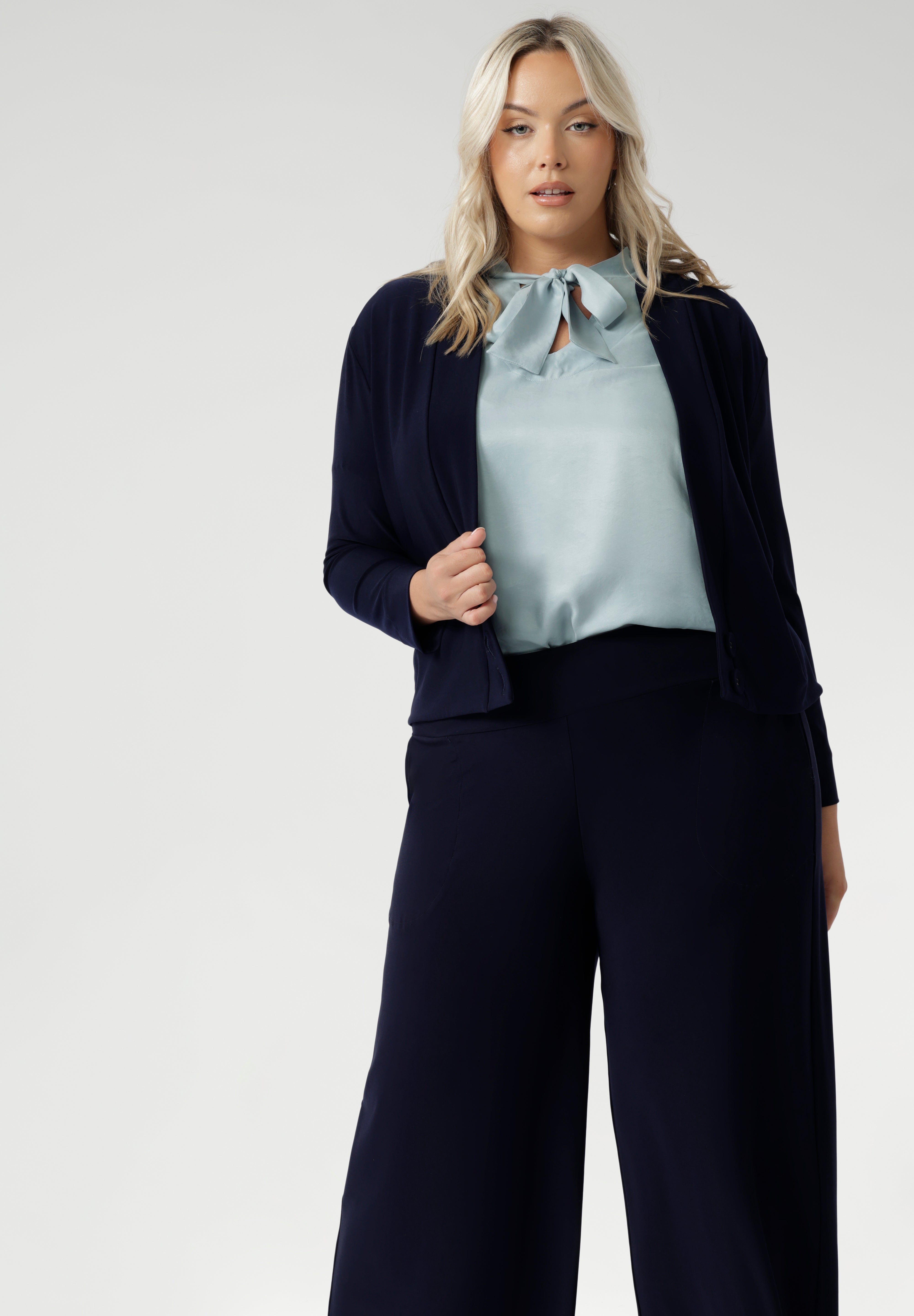 Plus size woman wears navy cardigan paired with woman's navy work pants and blue blouse with neck ties. The Jacardi features long sleeves, this jacket-cardigan hybrid is the perfect trans-seasonal cover up for women. Move into the cooler months in style, with this comfortable jacket for workwear and casual layering. Available in sizes 8-24.