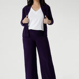 The Sandro Jacardi is a versatile jacket-like cardigan that effortlessly layers over dresses, making it an essential piece for any woman's workwear wardrobe. Petite height model wears purple jacket with matching purple wide leg pants and white top.