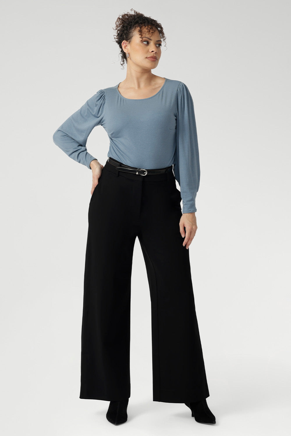 Size 10 Woman wears black wide leg work pants with a blue blouse tucked in and black boots. These black pants feature wide legs, belt loops, high waist and tailored details for excellent shaping. Available in sizes 8-24.
