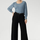 Size 10 Woman wears black wide leg work pants with a blue blouse tucked in and black boots. These black pants feature wide legs, belt loops, high waist and tailored details for excellent shaping. Available in sizes 8-24.