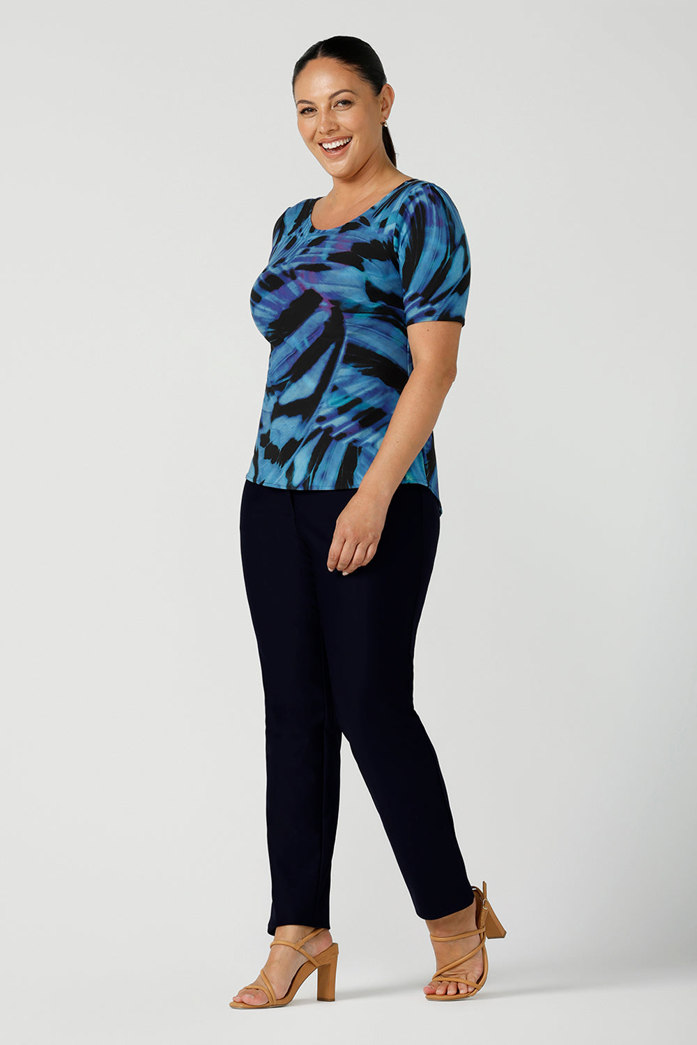 A size 12 woman wearing boat neckline Ziggy top in Flutter. A cobalt and purple print on a black base that is digitally printed. Made in Australia in soft Jersey with elbow length sleeves. A great work to weekend top. Size 8 - 24. Styled back with black slim fit Brooklyn pants and heels.