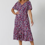 Summer dresses for over 50s women doen't get much beter than this! A size 12, over 50 woman wears an empire line midi dress with flutter sleeves and twist front bodice. A good dress for wedding guest outfits, the pink, red, white and navy blue print make it a pretty summer dress. Made in Australia by Australian and New Zealand women's clothes brand, Leina & Fleur, shop their petite to plus size dresses online. 