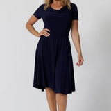 Summer dresses for over 40s women don't get much beter than this! A size 12, over 40 woman wears knee length summer dress with asymmetric hemlineand cowl neck. A short sleeve dress in navy jersey fabric, this is an easy-care dress and makes a good jersey dress for travel and cruise wear.  Made in Australia by Australian and New Zealand women's clothes brand, Leina & Fleur, shop their petite to plus size dresses online. 