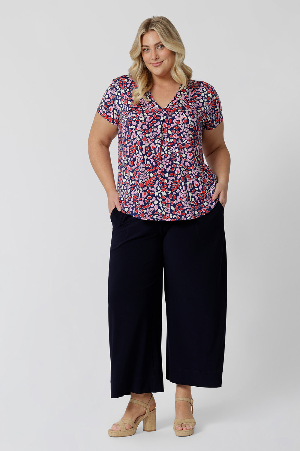 As an off-duty top or a work wear top for women, this V-neck top with tailroed tuck is a comfortable top for women.  A size 18 woman wears a a plus size short sleeve V-neck jersey top in red, pink, white and navy print with wide leg, navy pants. Made in Australia top by Australian and New Zealand women's clothes brand, Leina & Fleur, shop their petite to plus size tops online. 