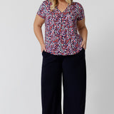 As an off-duty top or a work wear top for women, this V-neck top with tailroed tuck is a comfortable top for women.  A size 18 woman wears a a plus size short sleeve V-neck jersey top in red, pink, white and navy print with wide leg, navy pants. Made in Australia top by Australian and New Zealand women's clothes brand, Leina & Fleur, shop their petite to plus size tops online. 