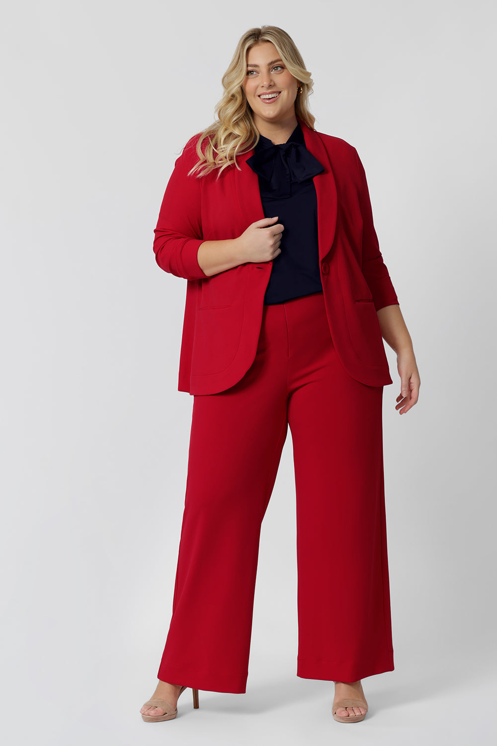 A size 18 woman wears the Merit Blazer in Flame, a textured scuba crepe plus size blazer with button front. This red work blazer is worn with red workwear pants and navy work top. Curved hemline and front pockets. This blazer is Made in Australia for curvy women size 8 - 24.