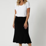 Over 40's woman wears black sleek skirt with streamlined design that can easily be dressed up for a more elevated look or worn casually - making it a truly versatile wardrobe staple. Model pairs black midid skirt with plain white top and a silver sneaker.