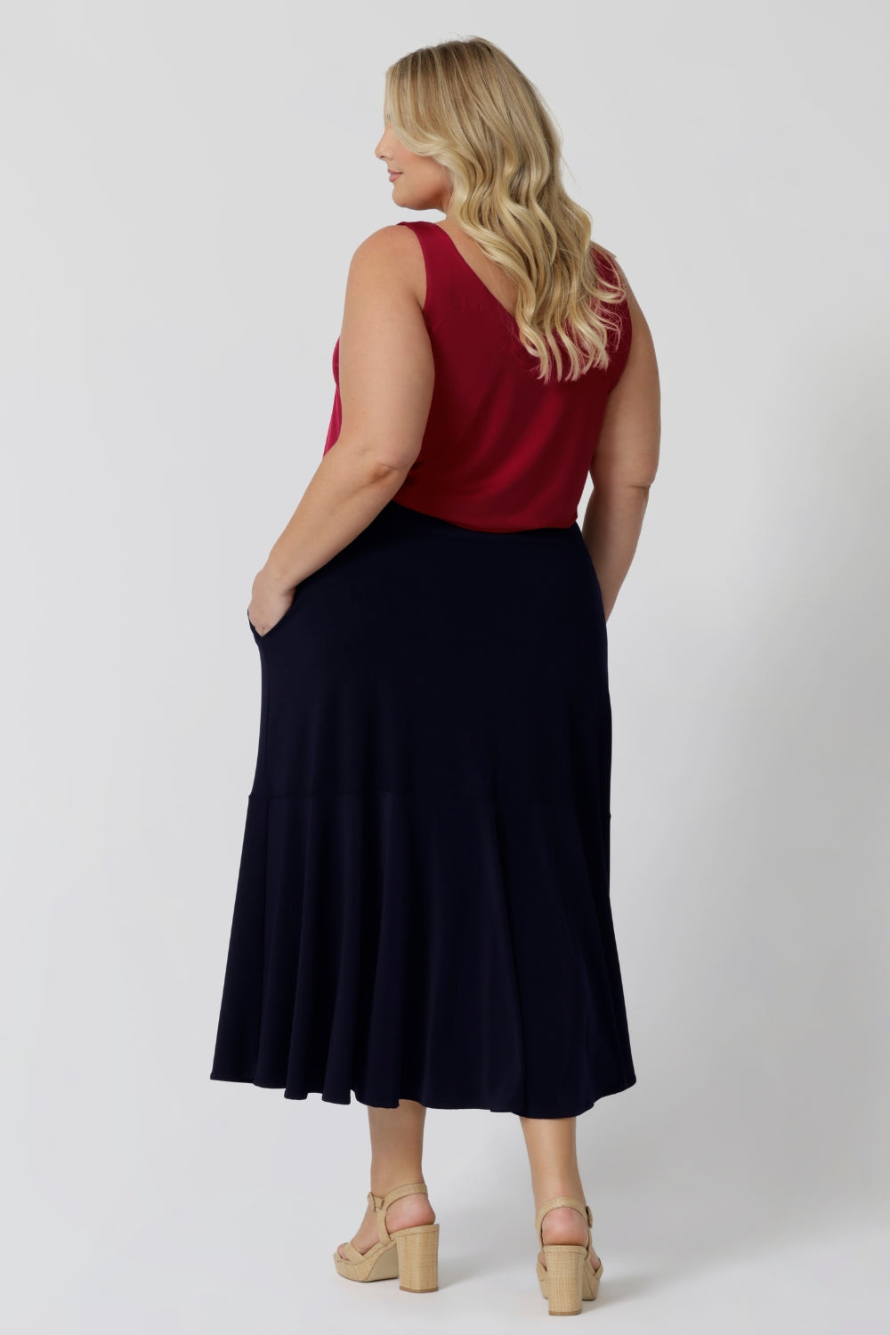 Back view of plus sized woman wears a pull on, navy maxi skirt with ruffle hemline worn with sleeveless cami top in red. This navy maxi skirt is a good workwear skirt or smart-casual skirt. Can be dressed up or down. Made in Australia in petite plus sizes for all curve of women's bodies. Berit Maxi Skirt in Navy available to shop in sizes 8-24. 