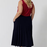 Back view of plus sized woman wears a pull on, navy maxi skirt with ruffle hemline worn with sleeveless cami top in red. This navy maxi skirt is a good workwear skirt or smart-casual skirt. Can be dressed up or down. Made in Australia in petite plus sizes for all curve of women's bodies. Berit Maxi Skirt in Navy available to shop in sizes 8-24. 