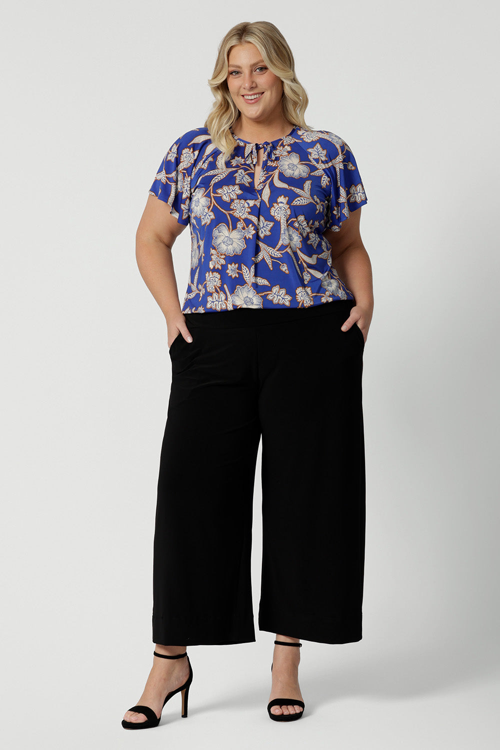 Wearing a good summer top for plus size women with flutter sleeves, a curvy size 18 woman shows the relaxed fit of a jersey top with wide leg black pants. With a holiday print and in lightweight, easy care jersey, this is the perfect top for travel and cruise wear. Made in Australia by Australian and New Zealand women's clothes label, Leina & Fleur shop plus size tops online.