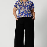 Wearing a good summer top for plus size women with flutter sleeves, a curvy size 18 woman shows the relaxed fit of a jersey top with wide leg black pants. With a holiday print and in lightweight, easy care jersey, this is the perfect top for travel and cruise wear. Made in Australia by Australian and New Zealand women's clothes label, Leina & Fleur shop plus size tops online.