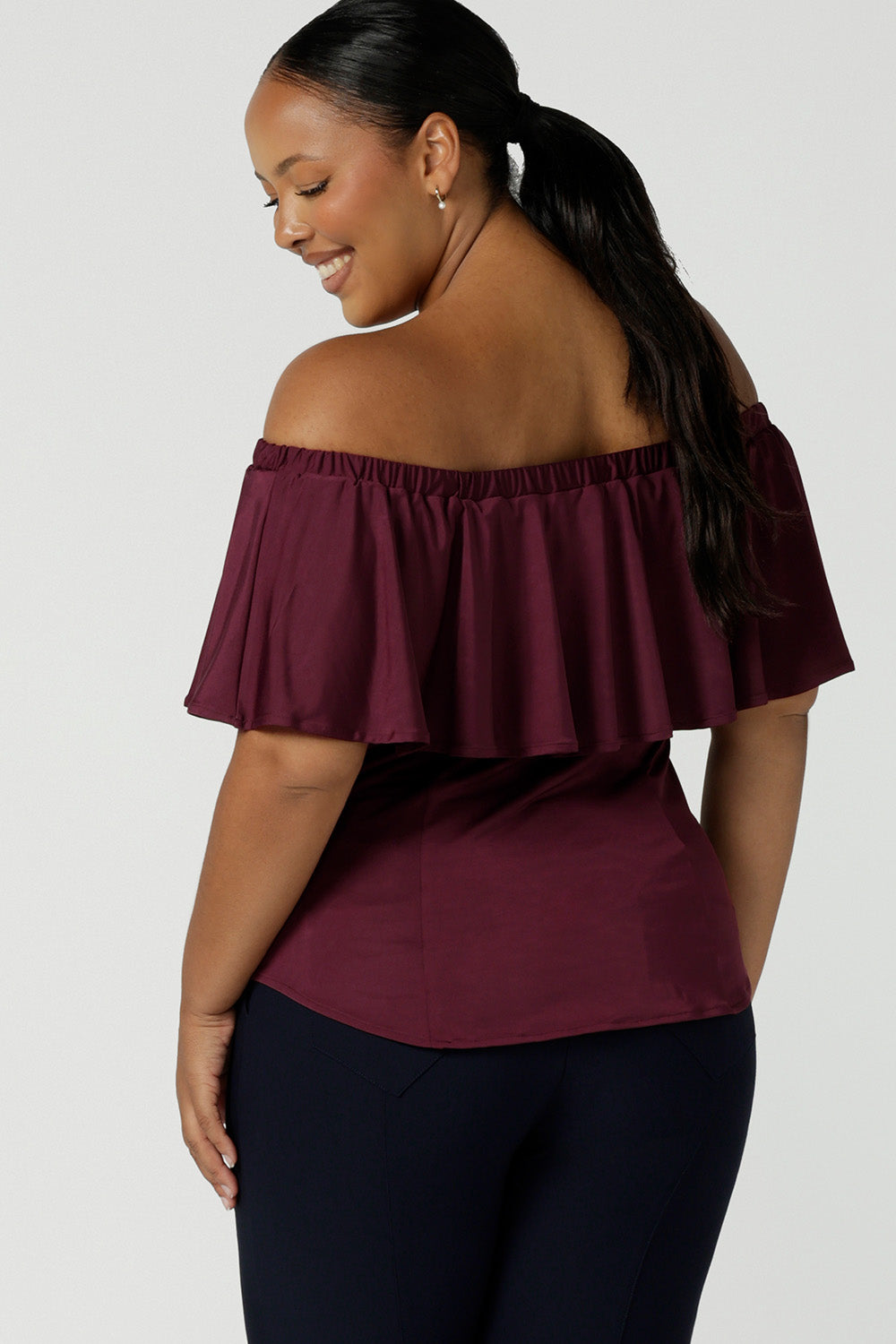 Back view of a plus size, size 18 woman wearing an off-the-shoulder top in plum slinky jersey. Worn with navy blue slim leg pants both are made in Australia by Australian and New Zealand women's clothing brand, Leina & Fleur. 