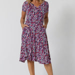 Summer dresses for over 40s women don't get much beter than this! A size 12, over 40 woman wears knee length summer dress with asymmetric hemlineand cowl neck. A short sleeve dress in pretty red, pink, white and navy print this is a good jersey dress for travel and cruise wear.  Made in Australia by Australian and New Zealand women's clothes brand, Leina & Fleur, shop their petite to plus size dresses online. 