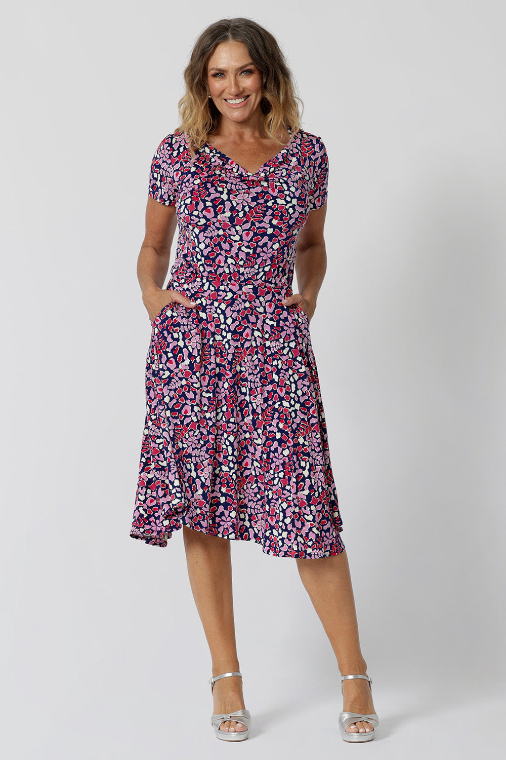 Summer dresses for over 40s women don't get much beter than this! A size 12, over 40 woman wears knee length summer dress with asymmetric hemlineand cowl neck. A short sleeve dress in pretty red, pink, white and navy print this is a good jersey dress for travel and cruise wear.  Made in Australia by Australian and New Zealand women's clothes brand, Leina & Fleur, shop their petite to plus size dresses online. 