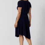 Back view of a navy jersey dress for over 40s women. A size 12, over 40 woman wears knee length summer dress with asymmetric hemlineand cowl neck. A short sleeve dress in navy jersey fabric, this is an easy-care dress and makes a good jersey dress for travel and cruise wear.  Made in Australia by Australian and New Zealand women's clothes brand, Leina & Fleur, shop their petite to plus size dresses online. 
