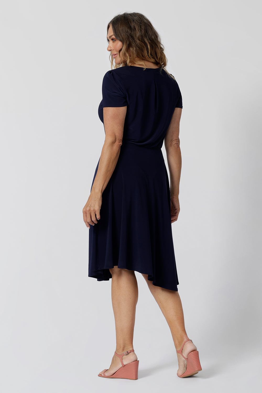 Back view of a navy jersey dress for over 40s women. A size 12, over 40 woman wears knee length summer dress with asymmetric hemlineand cowl neck. A short sleeve dress in navy jersey fabric, this is an easy-care dress and makes a good jersey dress for travel and cruise wear.  Made in Australia by Australian and New Zealand women's clothes brand, Leina & Fleur, shop their petite to plus size dresses online. 