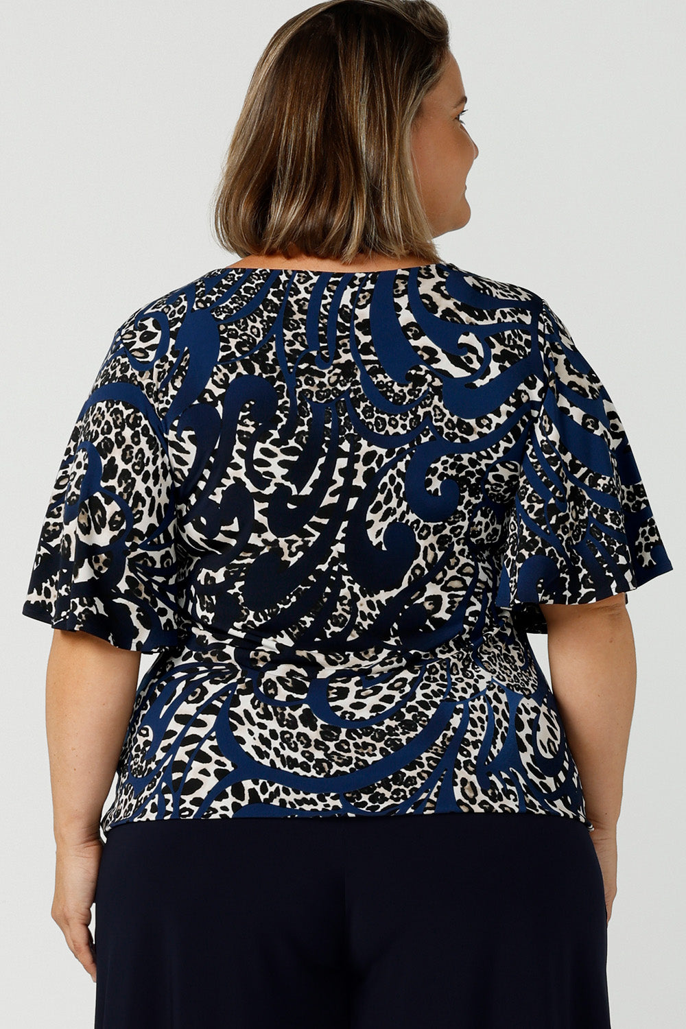 Back view of a petite height size 16 woman wears a jersey top in animal print, styled with navy wide leg pants. This Australian-made women's top has flutter sleeves, a high, round neckline and and high-low hem perfect for weekend casual and travel capsule wardrobes.