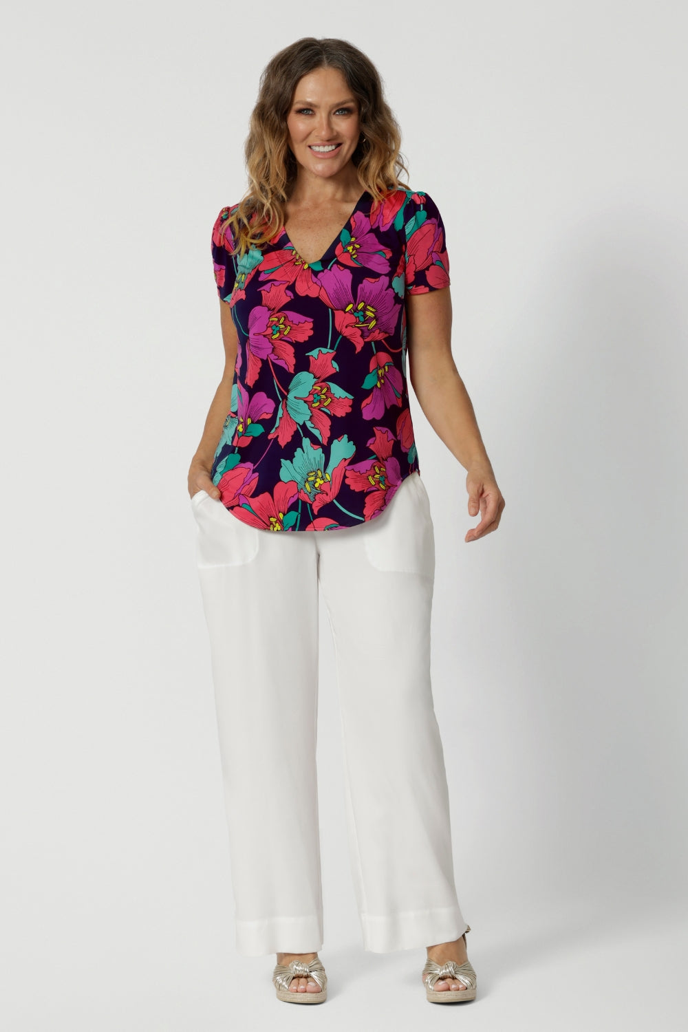 A curvy size 10 woman over 40 wears a top with a v-neckline and short sleeves. It has a bold floral print. She has styled the top with white resort pants. The clothes are handmade in Australia by Australian fashion brand Leina & Fleur in sizes 8 to 24. 