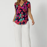 A curvy size 10 woman over 40 wears a top with a v-neckline and short sleeves. It has a bold floral print. She has styled the top with white resort pants. The clothes are handmade in Australia by Australian fashion brand Leina & Fleur in sizes 8 to 24. 