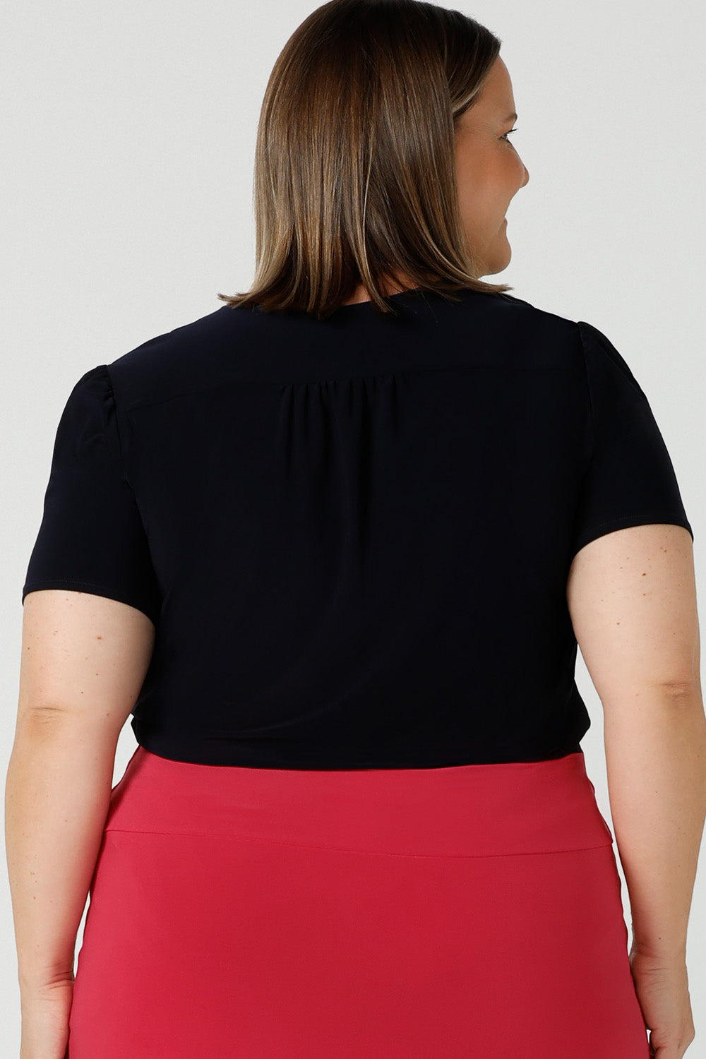  Back view of a curvy size 18 woman wears a navy jersey top great for corporate wear.  Styled back with a fuchsia pink tube skirt. This tailored top with short sleeves and a v-neckline is made in Australia by Australian fashion brand, Leina & Fleur in sizes 8 to 24.